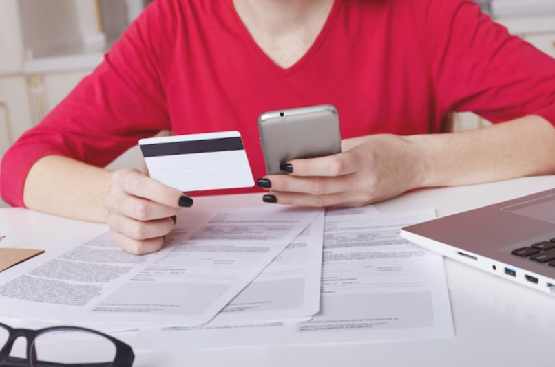 Unrecognizable female model in red sweater sits at working table surrounded with papers and laptop computer
