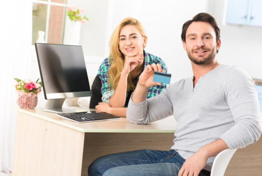 toned of happy and cheerful young man showing credit card to camera. man and woman smiling for camera while working in office interior.